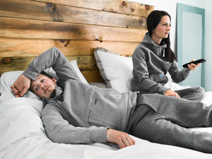 2 Tuxy Users Laying in bed. One male user wearing a grey tuxy suit is laying with head on pillow. Other female tuxy user is sitting up in grey suit holding TV remote 
